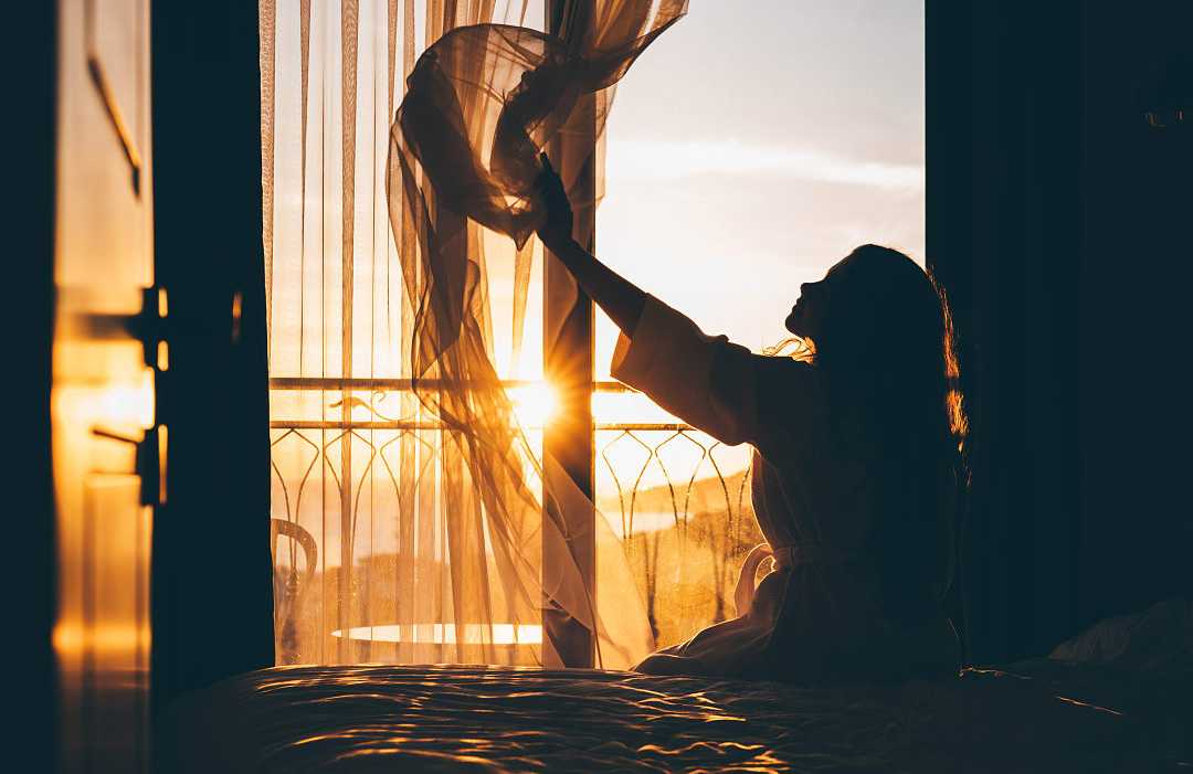 Woman sitting on a bed and opening curtains as the morning sunlight streams into a hotel room.