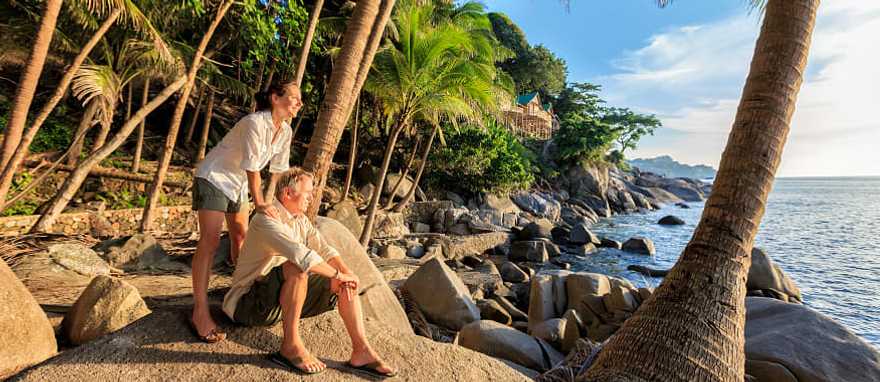 Senior couple enjoying a tropical beach at sunset surrounded by palm trees and rocky shoreline in Thailand.