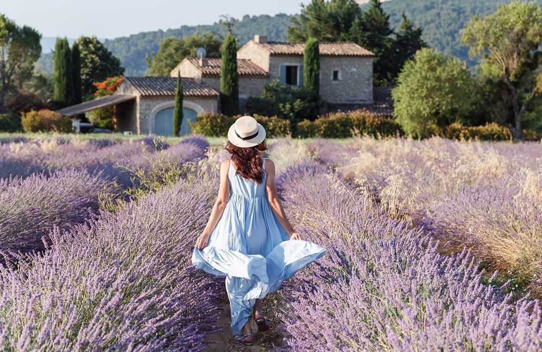 Woman in blue dress walking through a lavender field in Luberon, France, with rustic stone house behind