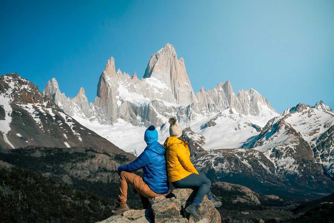 wo hikers in colorful jackets admire snowy Mount Fitz Roy in Patagonia under a clear blue sky