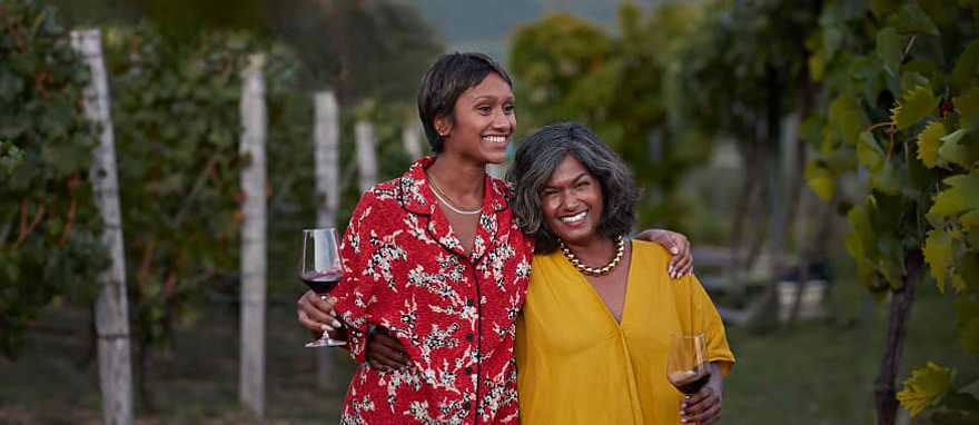 Smiling mother and daughter holding wineglasses and standing with arms around each other at a vineyard