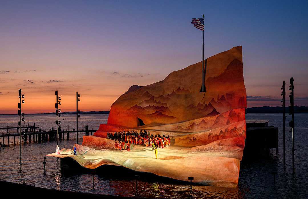 Open-air opera performance on a floating stage at sunset with a dramatic backdrop and calm waters