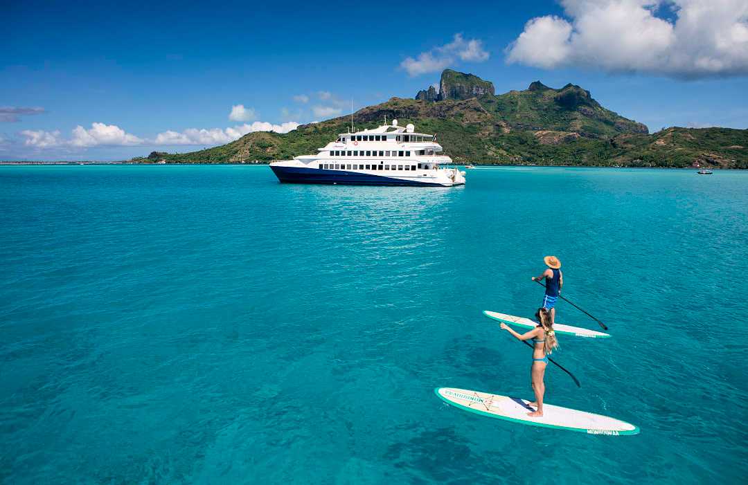 Couple paddleboarding on turquoise water in Bora Bora with a luxury yacht and tropical island backdrop