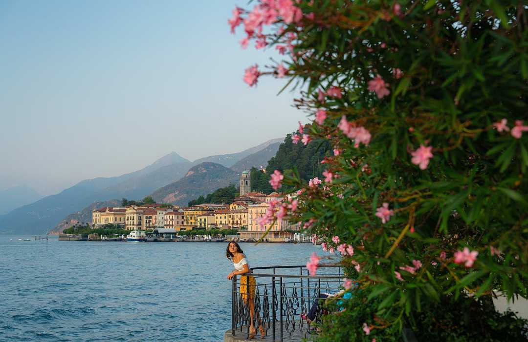 Scenic view of a woman by Lake Como, vibrant flowers, waterfront architecture, and mountains in the distance