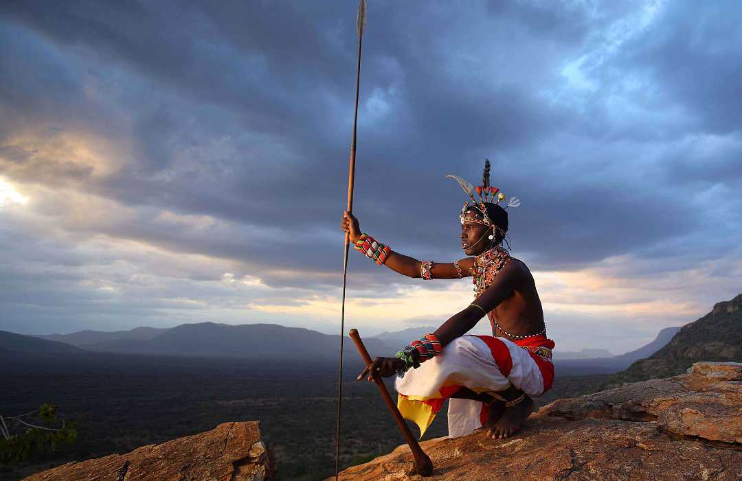 Kenya, Africa. Photo courtesy of: Kamala Conservancy A traditional warrior sitting on a rocky hill at sunset, holding a spear and overlooking a vast wilderness landscape.